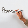 Ein Hand schreibt mit einem blauen Marker das Wort "Planen" auf eine weiße Oberfläche.