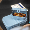 Blaue Lunchbox mit Nüssen auf einem Tisch, weitere Box darunter, Notizblock im Hintergrund.