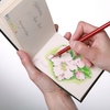 Offene Seiten eines Skizzenbuchs mit einer Hand, die mit einem Farbstift auf eine Blume zeichnet.