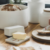 Weiße Butterdose mit Butterstück auf einem Tisch neben Brot und dekorativen Behältern.