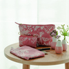 Rosa Kosmetiktasche mit floralem Muster auf einem Tisch, umgeben von weiteren Taschen und Vasen.