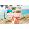 Weißer Füllfederhalter liegt auf Postkarte mit Urlaubsfotos in einer Hand vor einer Strandkulisse.