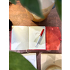 Rotes Notizbuch geöffnet mit leeren Seiten und einem Schreibgerät auf einem Tisch unter Pflanzen.