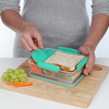 Hand, die eine sistema Lunchbox öffnet, auf einem Holzbrett mit Trauben und Karotten daneben.