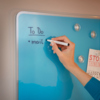 Blaue Glasoberfläche eines Whiteboards mit handschriftlicher Notiz und Magneten.