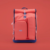 Rosa Schulrucksack mit zwei Seitentaschen und Rolltop-Verschluss auf rotem Hintergrund.