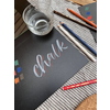 Buntstifte auf einem Tisch mit einem Glas und einer Kreideinschrift auf schwarzem Papier.