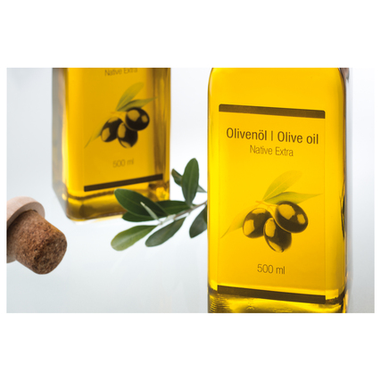Transparente Flasche mit Olivenöl und Etikett, neben einer Korken und Olivenzweig.