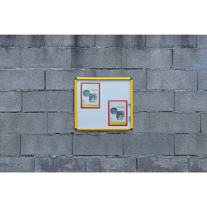 Schaukasten mit gelbem Rahmen an einer grauen Betonwand, mit zwei DIN A4-Dokumenten darin.
