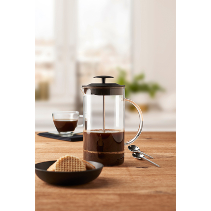 Kaffeebereiter aus Glas mit schwarzem Deckel, neben einer Tasse Kaffee und Waffeln auf einem Tisch.