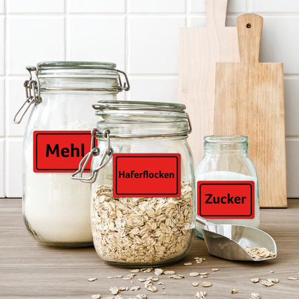 Neonrote Etiketten auf Gläsern mit Mehl, Haferflocken und Zucker auf einem Holztisch.