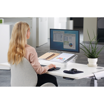 Frau sitzt an einem Schreibtisch mit Monitor, Dokumentenhalter und Pflanzen im Hintergrund.