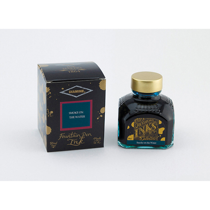 Glasflasche mit schwarzer Tinte und goldfarbenem Deckel neben schwarzer Verpackung.