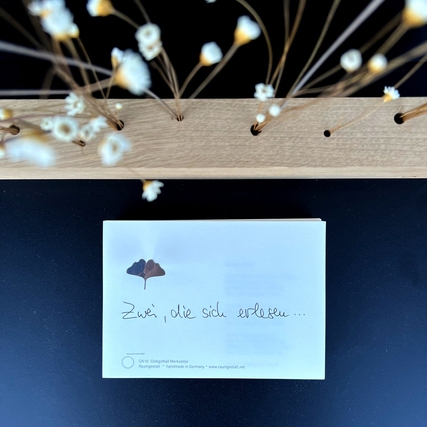 Notizblock mit Ginkgo-Illustration auf Eichenklotz, dekoriert mit Trockenblumen im Hintergrund.