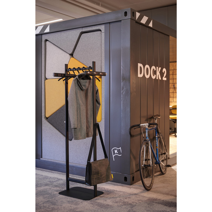 Stehende Garderobe mit Kleidungsstücken und Tasche, platziert vor farbiger Wand mit Fahrrad.