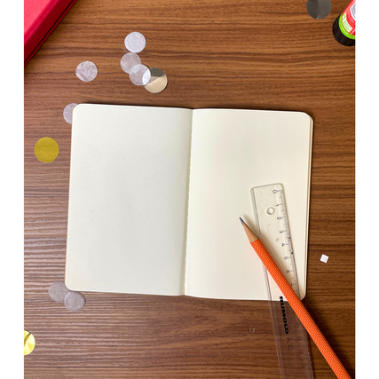 Offenes Notizheft auf einem Tisch mit Bleistift und Lineal, umgeben von bunten Bastelmaterialien.