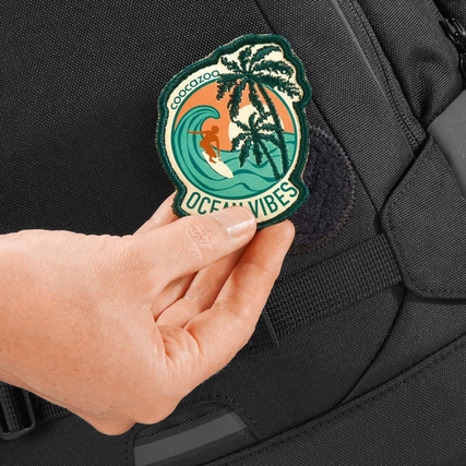 Person hält ein farbigen Patch mit "Ocean Vibes" an einem schwarzen Rucksack.