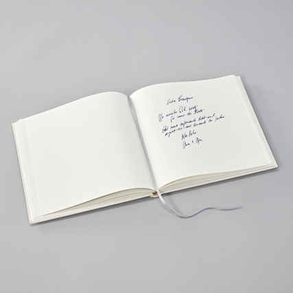 Offenes Gästebuch mit leeren Seiten und handschriftlichem Text auf grauem Untergrund.