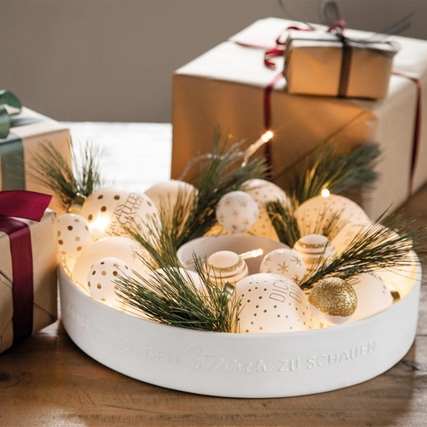 Runder Kerzenständer mit dekorativen Elementen und Weihnachtsgeschenken im Hintergrund.