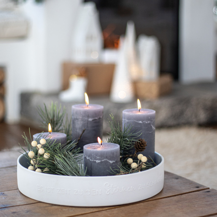 Weißer Kerzenständer mit vier dekorativen Kerzen, umgeben von grünen Zweigen und Beeren.