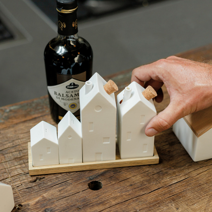Weiße Porzellanhäuser auf Holzunterlage mit einer Hand, neben einer Flasche Balsamico.