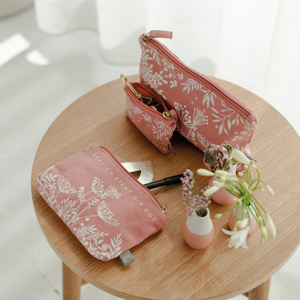 Zwei rosa Kosmetiktaschen mit floralem Muster und Inhalt auf einem Tisch, dekoriert mit Blumen.