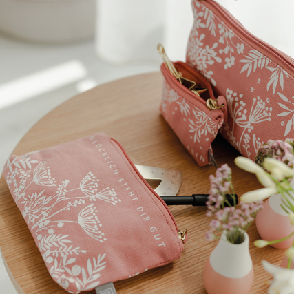 Rosa Kosmetiktasche mit floralem Muster und Schriftzug, auf einem Tisch mit Dekoration.