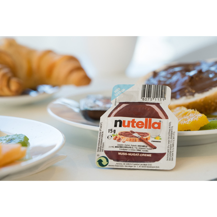 Ein Portionsbecher Nutella Nuss-Nugat-Creme auf einem Tisch mit Croissants und Obst.