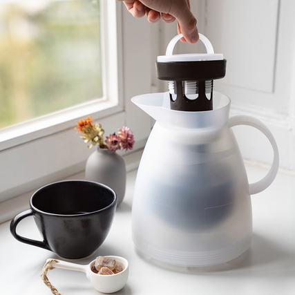Isolierkanne mit Teeeinsatz und Tasse auf einem Tisch, mit Blumen im Hintergrund.