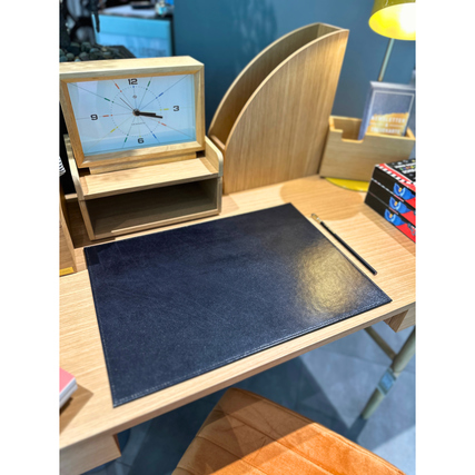 Schwarze Schreibunterlage auf einem Schreibtisch mit Uhr, Lampe und Büchern im Hintergrund.
