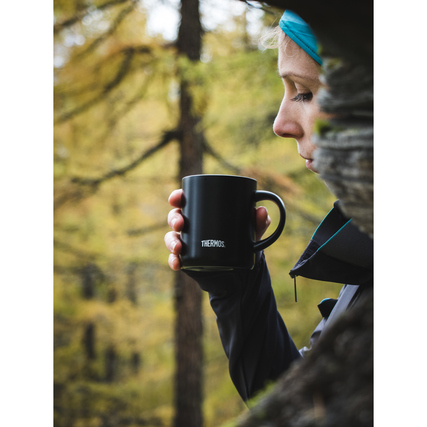 Schwarzer Thermobecher in Hand eines Benutzers, umgeben von einer herbstlichen Waldlandschaft.