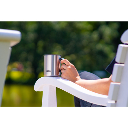 Edelstahl-Thermobecher in Hand einer Person auf einer weißen Gartenliege am Wasser.