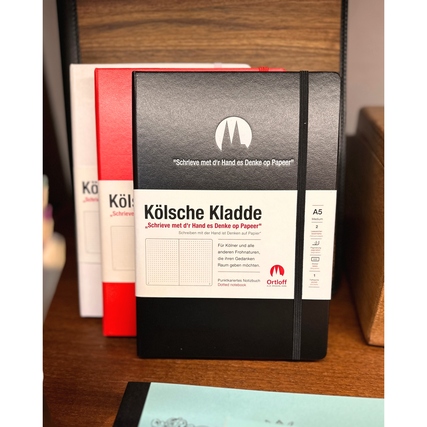 Schwarzes DIN A5 Notizbuch mit "Kölsche Kladde"-Prägung und Gummiband auf einem Holzuntergrund.