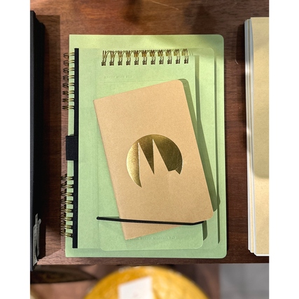 Kleines Notizheft in Kraftpapier mit goldener Logo-Prägung auf einem Tisch mit weiteren Heften.