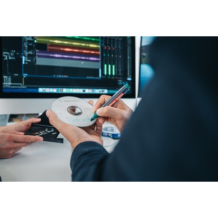 Person schreibt mit einem Marker auf eine CD-R, die auf einem Tisch neben einem Computer liegt.