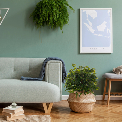 Künstliche Zimmerpflanze Ficus II in einem dekorativen Korb auf einem Holzboden neben einer Couch.