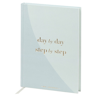 Hellblaues Notizbuch mit goldfarbener Prägung "day by day, step by step" auf dem Cover.