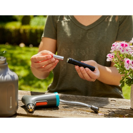 Person hält die Batterie und einen Teil der GARDENA Sprühflasche EasyPump in einer Gartenumgebung.