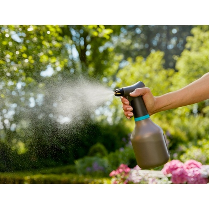 Pumpsprüher mit Wassernebel in einer Hand, im Garten mit Blumen im Hintergrund.
