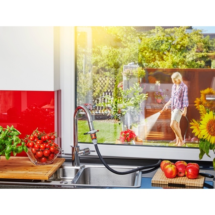 Gartenbrause im Einsatz zur Bewässerung von Pflanzen, mit Küche und Garten im Hintergrund.