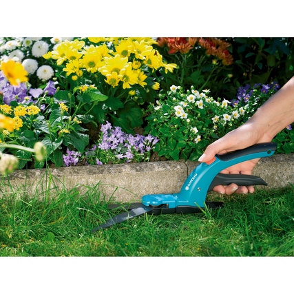 Hand mit GARDENA Grasschere schneidet Rasenkanten vor bunten Gartenblumen.