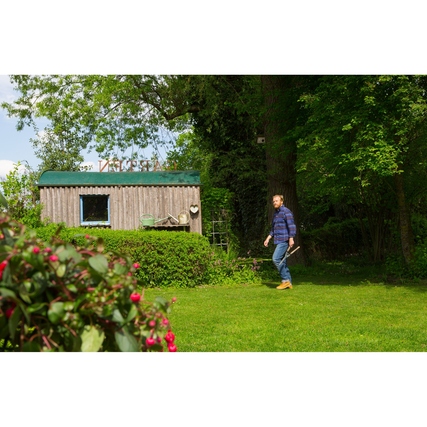 Mann mit Heckenschere geht auf gepflegter Rasenfläche neben Gartenhaus und Büschen.