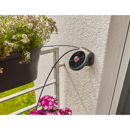 Bewässerungssystem mit Halterung an einer Wand und Wasserrohren, umgeben von blühenden Pflanzen.