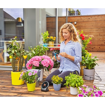 Frau mit Bewässerungssystem zwischen blühenden Pflanzen auf einer Terrasse.