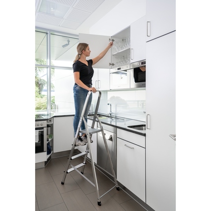 Frau erreicht Gläser in einem Küchenschrank mit einer einseitig begehbaren Stehleiter.