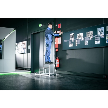 Mann steht auf einer Aluminium-Podestleiter und erreicht eine Wand in einem modernen Raum.
