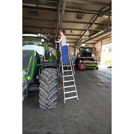 Person steht auf einer Stehleiter und arbeitet an einem Traktor in einer Werkstatt.