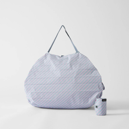 Shupatto Tragetasche L in Hellblau mit pinken Streifen, neben einer kleinen, gerollten Tasche.