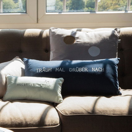 Zwei verschiedene Kissen mit Text und Mustern auf einer Couch in einem hellen Raum.