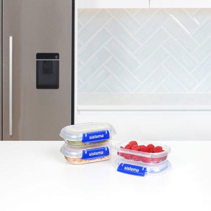 Drei transparente Vorratsdosen mit Snacks und Früchten auf einem Tisch vor einem Kühlschrank.